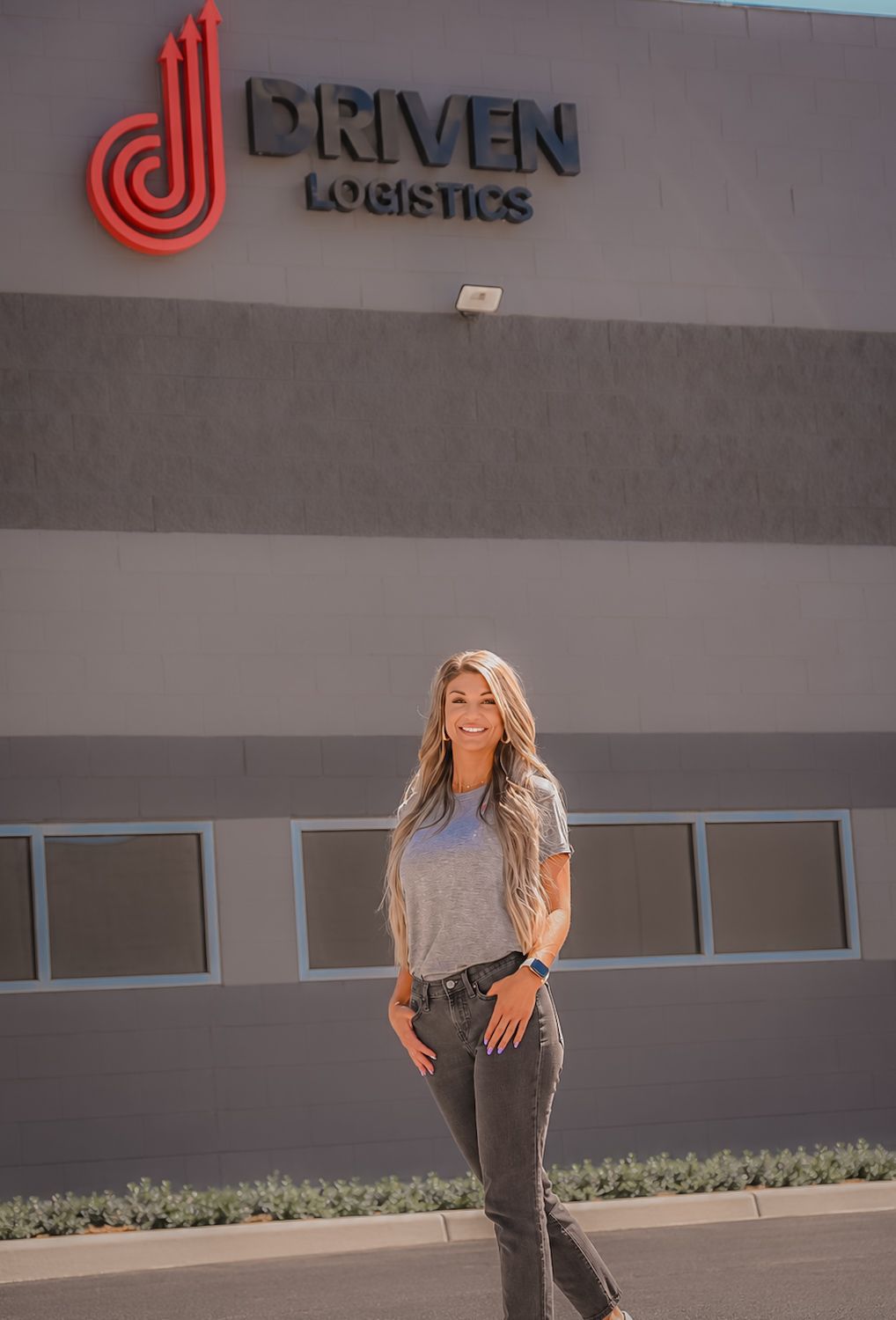 Woman smiling in front of Driven Logistics building exterior sign in sunny outdoor setting.