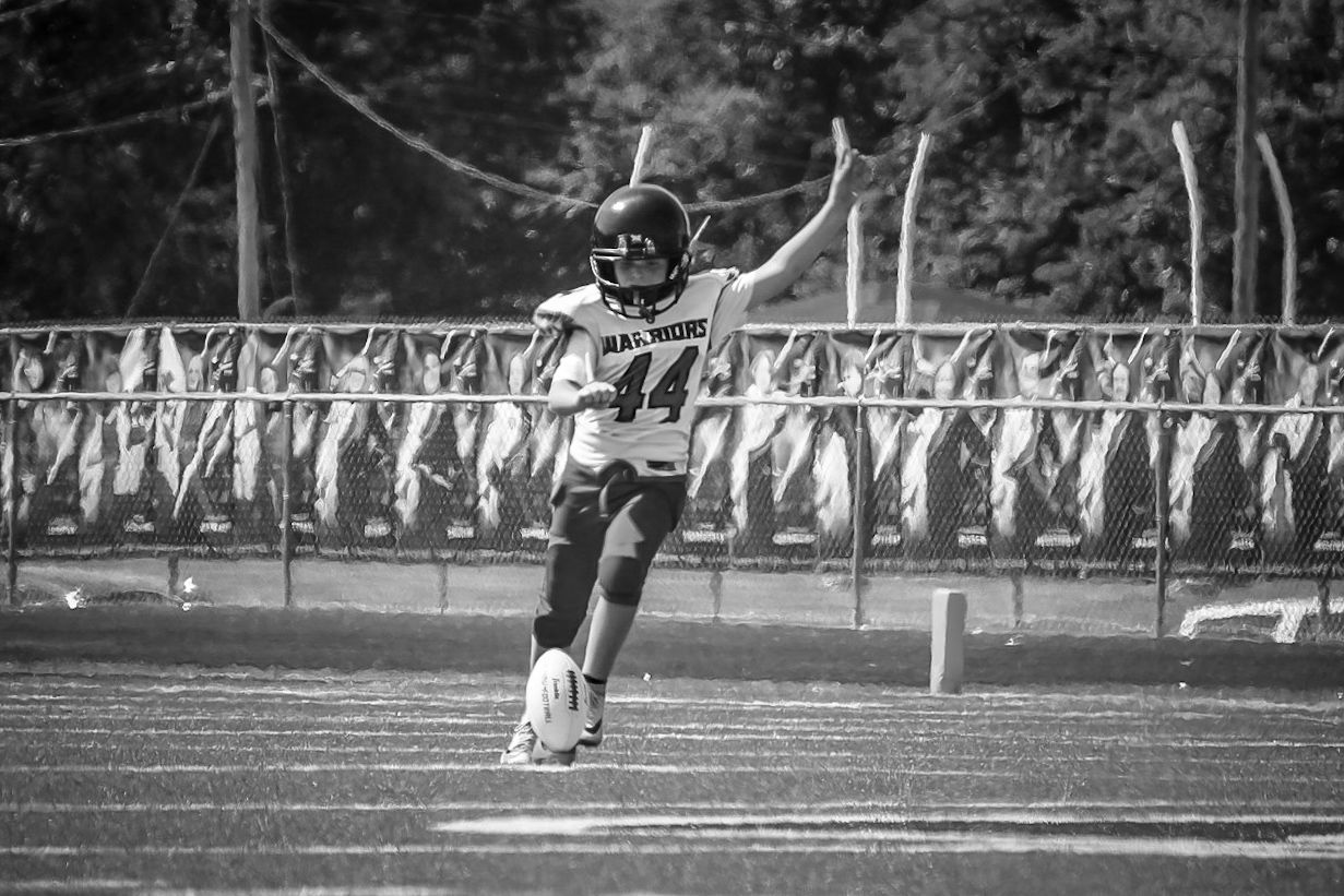 Black and white photo of a football player mid-kick on a field during a game.
