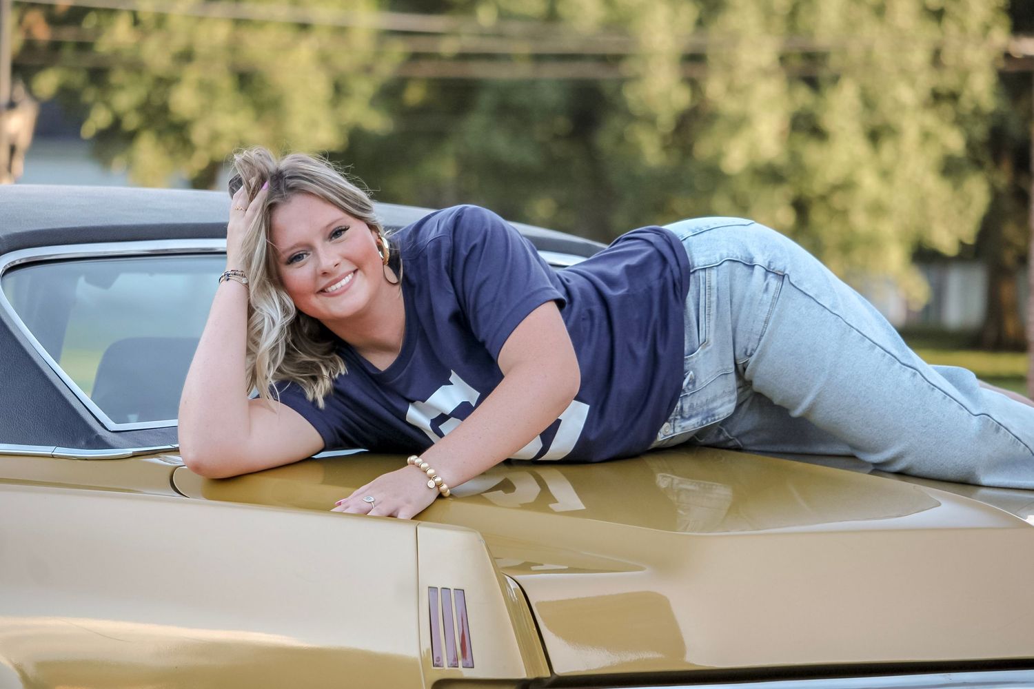 Smiling blonde woman posing on the hood of a classic gold muscle car outdoors.