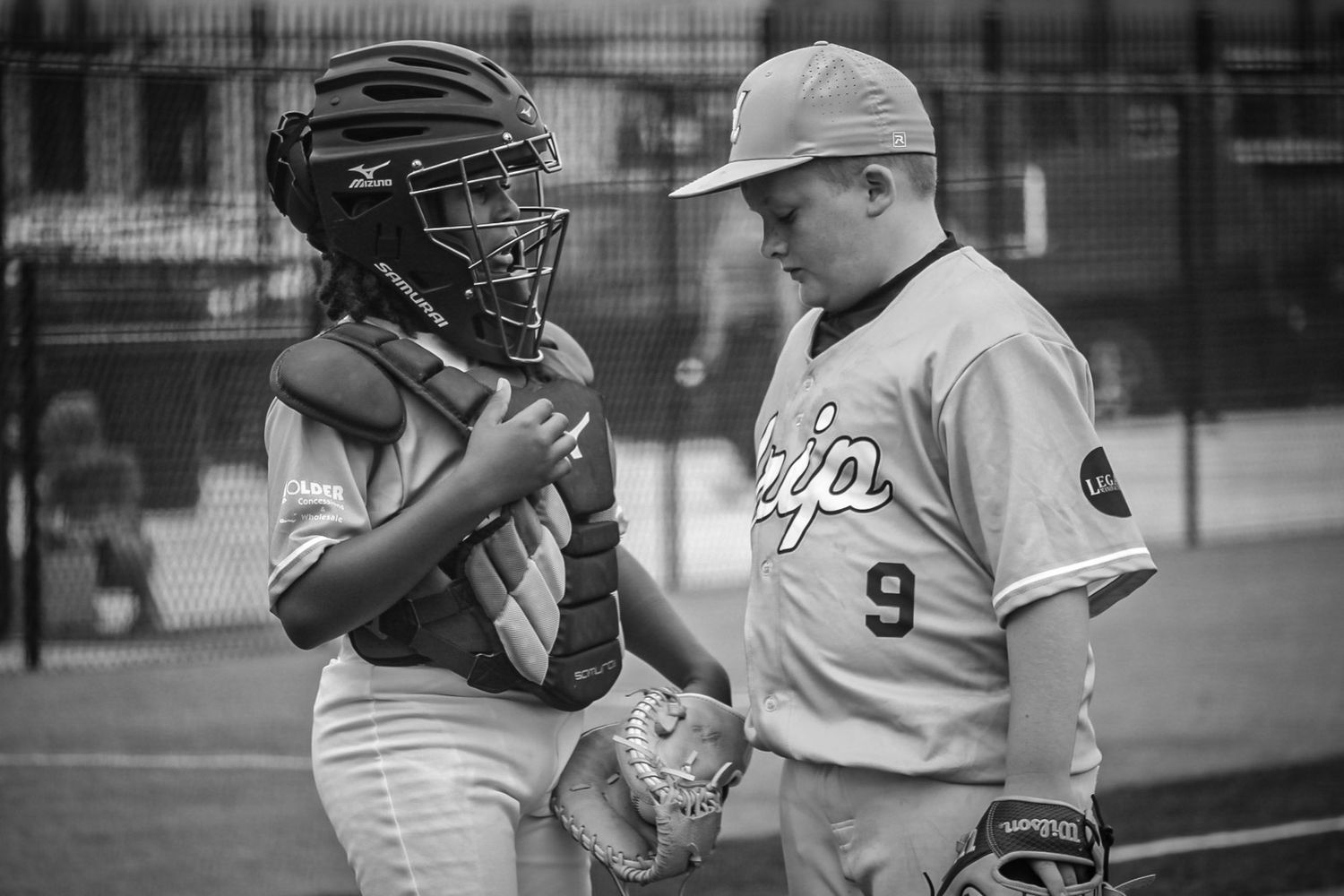 Two youth baseball players in uniform having a discussion on the field, one wearing catcher gear.