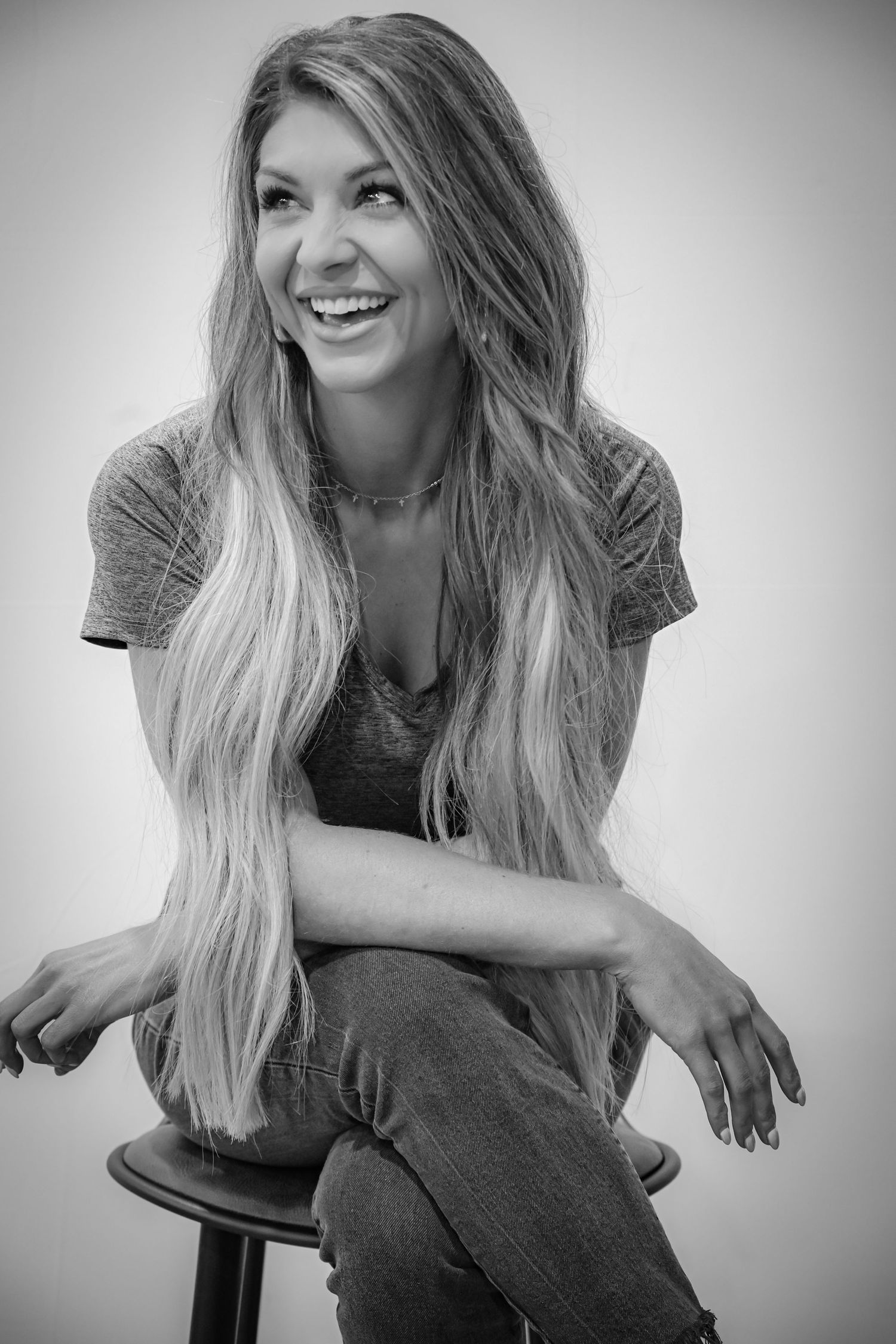 Black and white portrait of smiling woman with long blonde hair sitting on stool laughing.