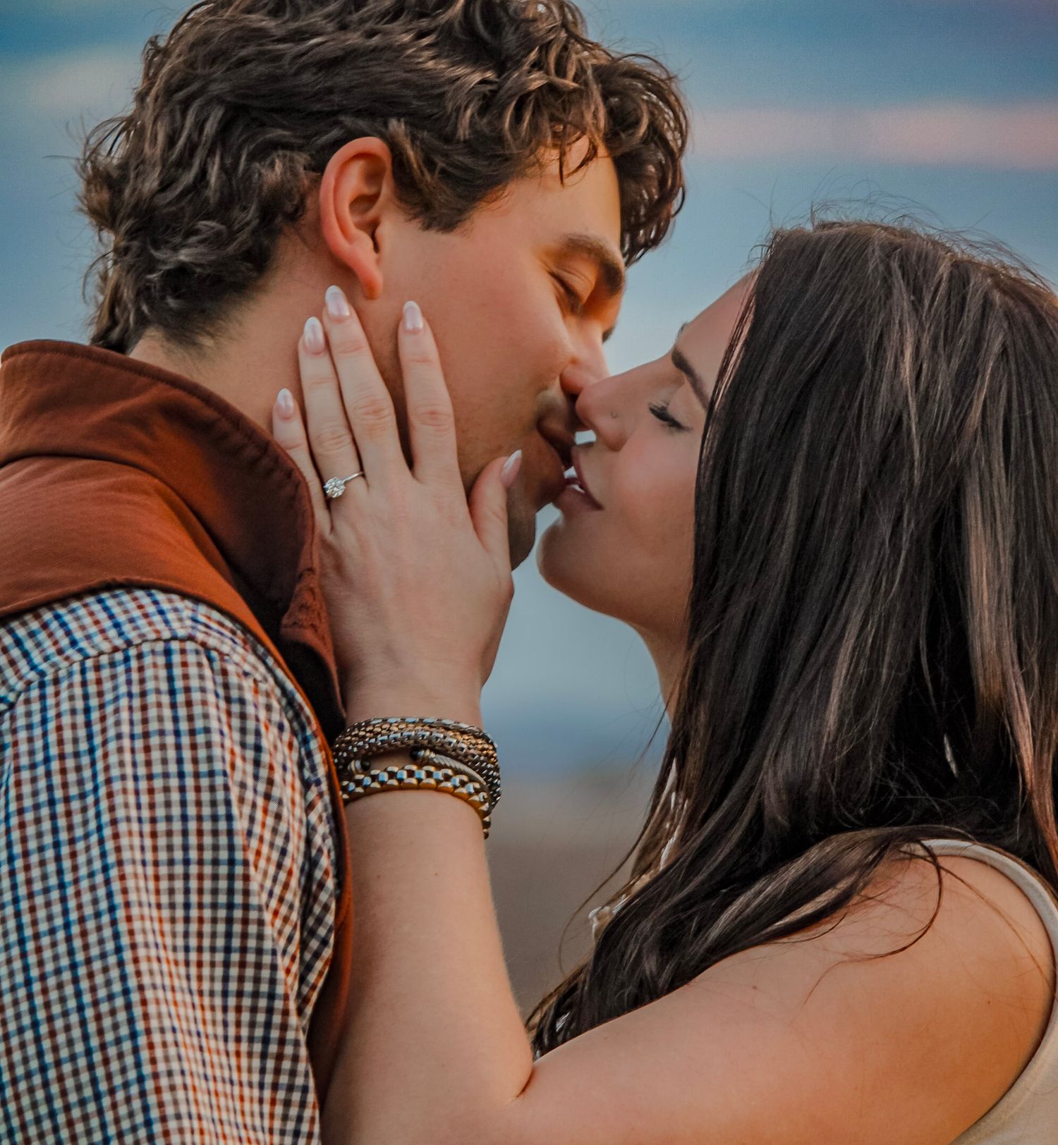 Couple sharing a romantic kiss outdoors at sunset, woman wearing an engagement ring.