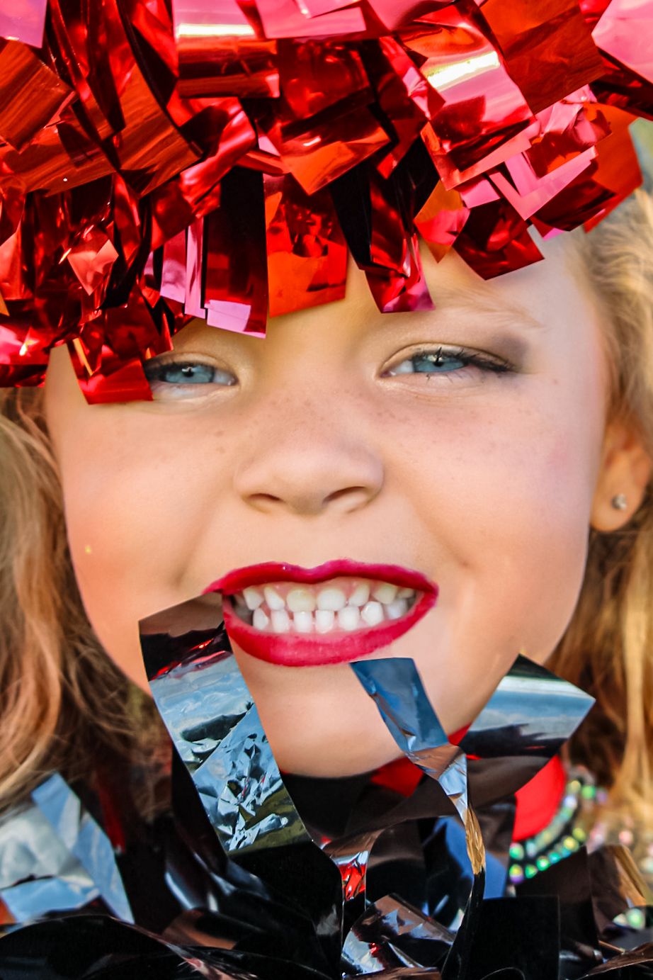 Young girl with red lipstick smiling, covered in shiny red and silver metallic confetti streamers.