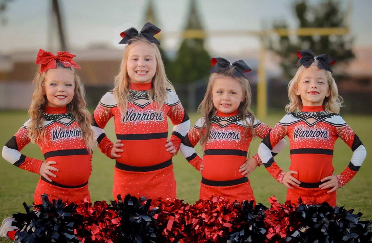 Four young girls in red Warriors cheerleader uniforms posing with pom poms on a field.