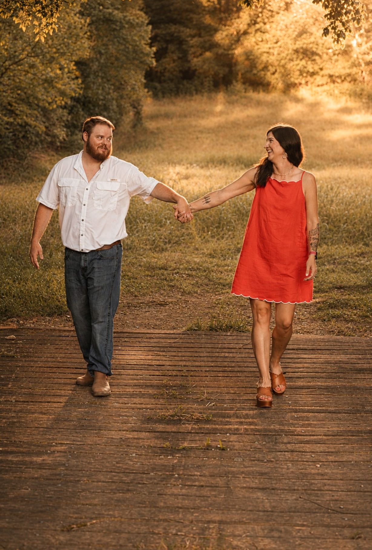Couple holding hands walking on a wooden path surrounded by lush green trees in golden hour light.