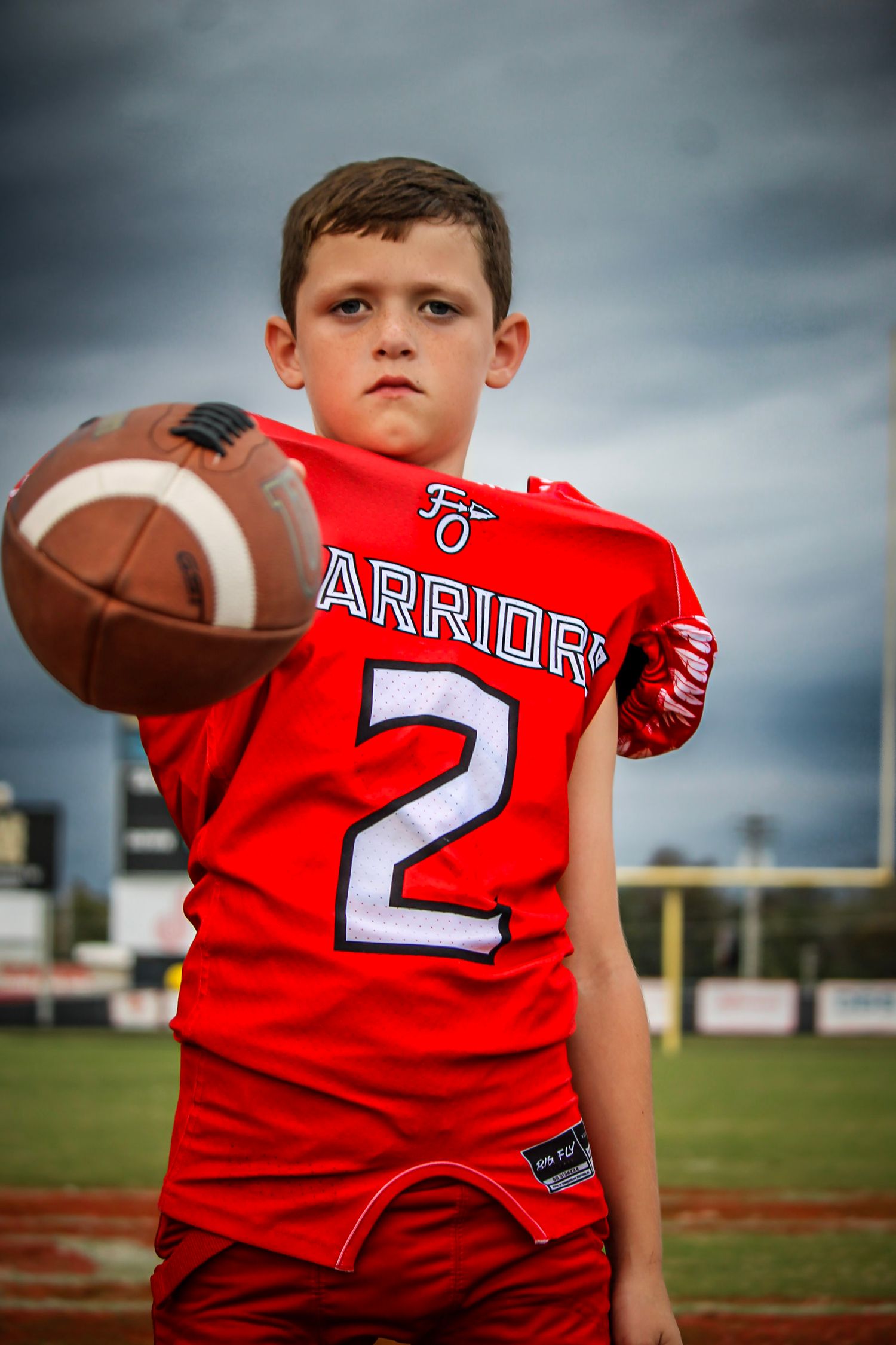 Young Warriors football player in red jersey number 2 holding a football toward camera.