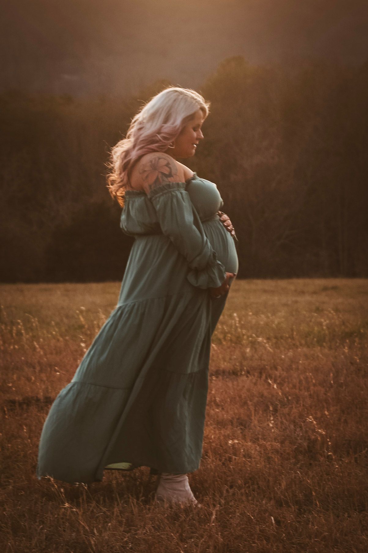 Pregnant woman in teal dress cradling baby bump in a sunlit golden field at sunset.