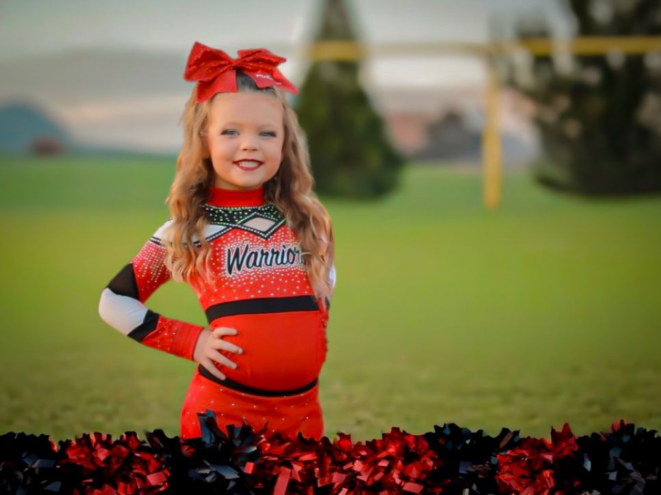Young girl in red Warriors cheerleader uniform with red bow and pom poms posing on green field.