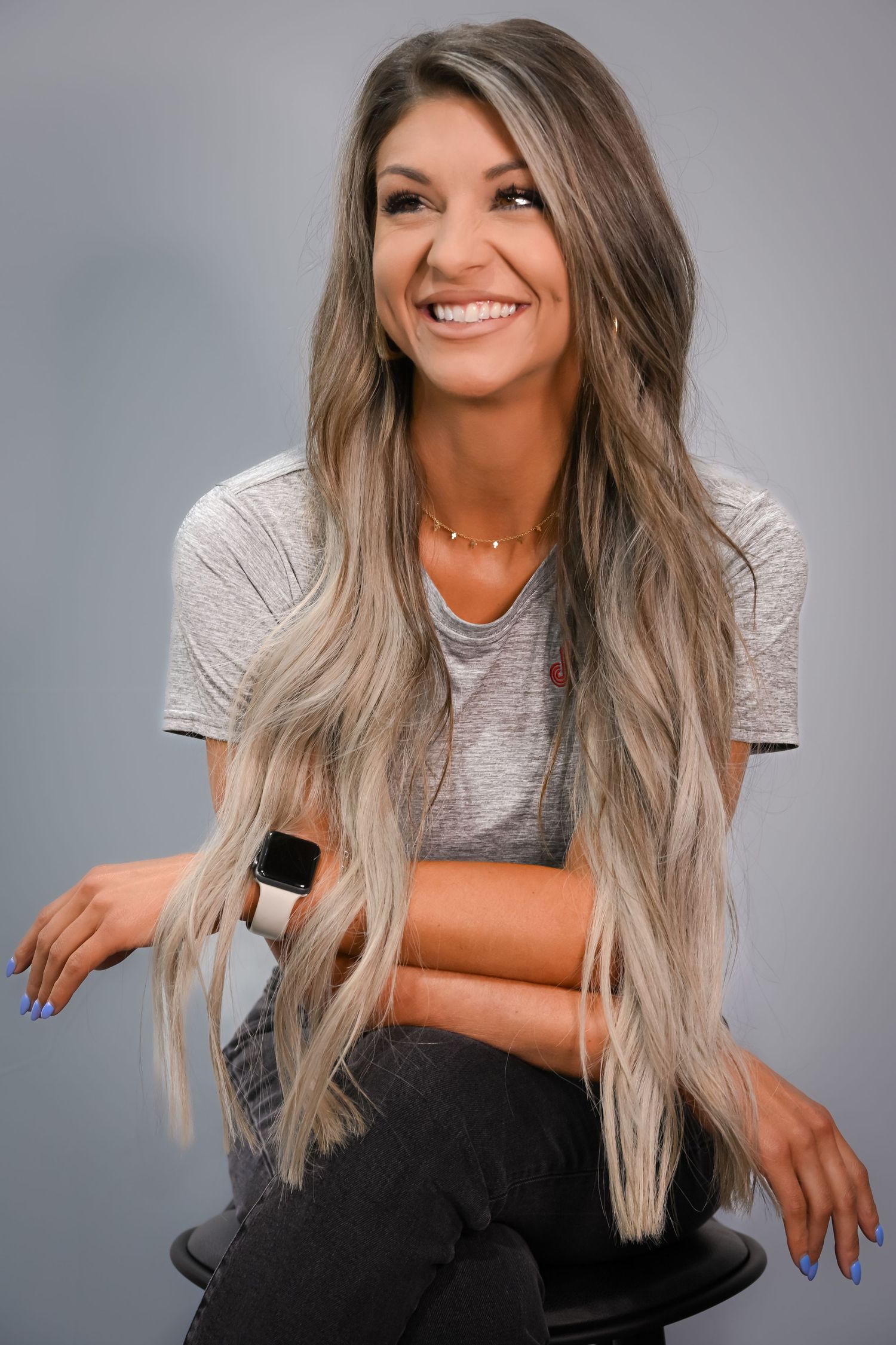 Smiling woman with long blonde highlighted hair sitting on stool wearing gray t-shirt and black jeans.