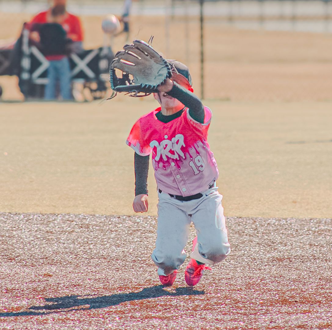 Young baseball player in pink jersey catching a ball with glove raised during a youth league game on a dirt field.
