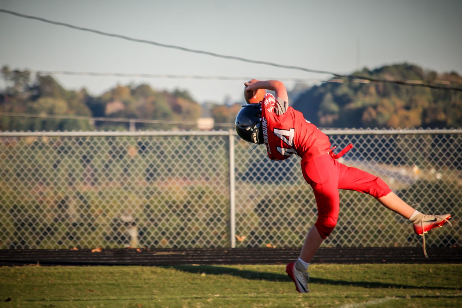 Football player in red uniform leaping to catch a pass during a sunny outdoor game near a chain-link fence.