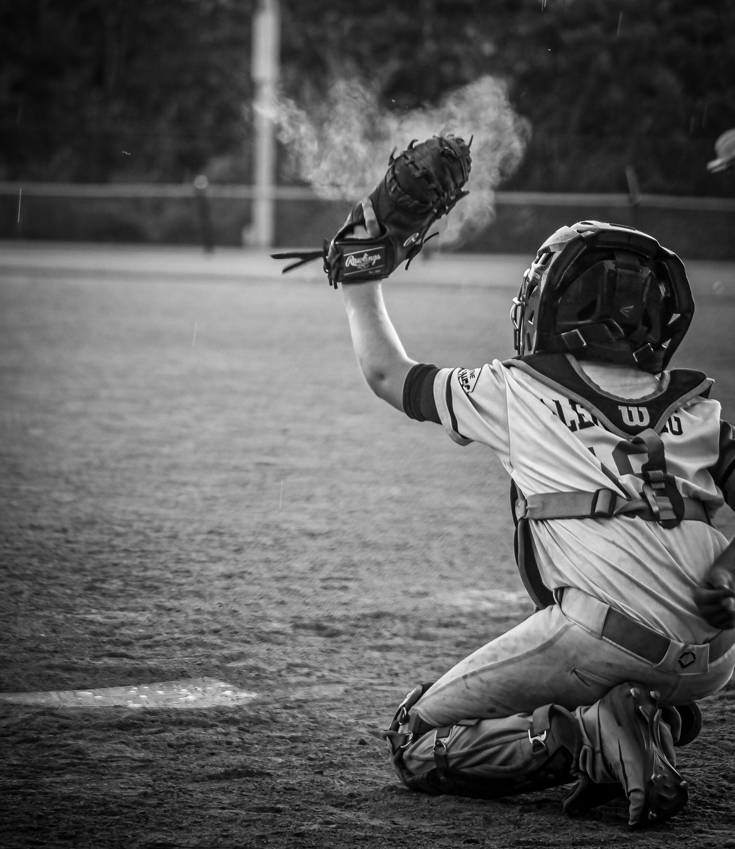 Black and white photo of a baseball catcher in full gear squatting and catching a pitch with visible breath vapor.
