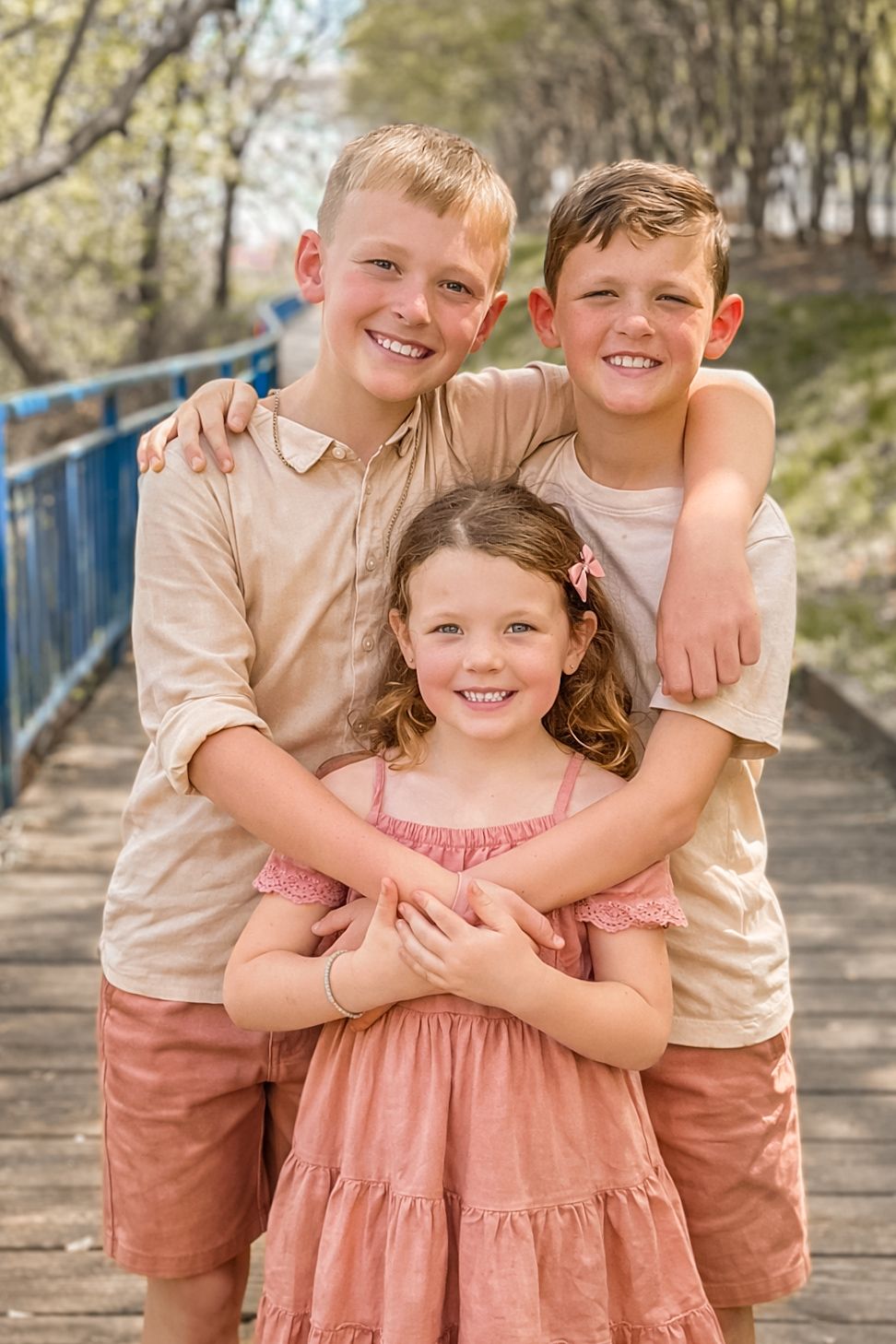 Three smiling children posing together on a wooden bridge outdoors in coordinating beige and pink outfits.