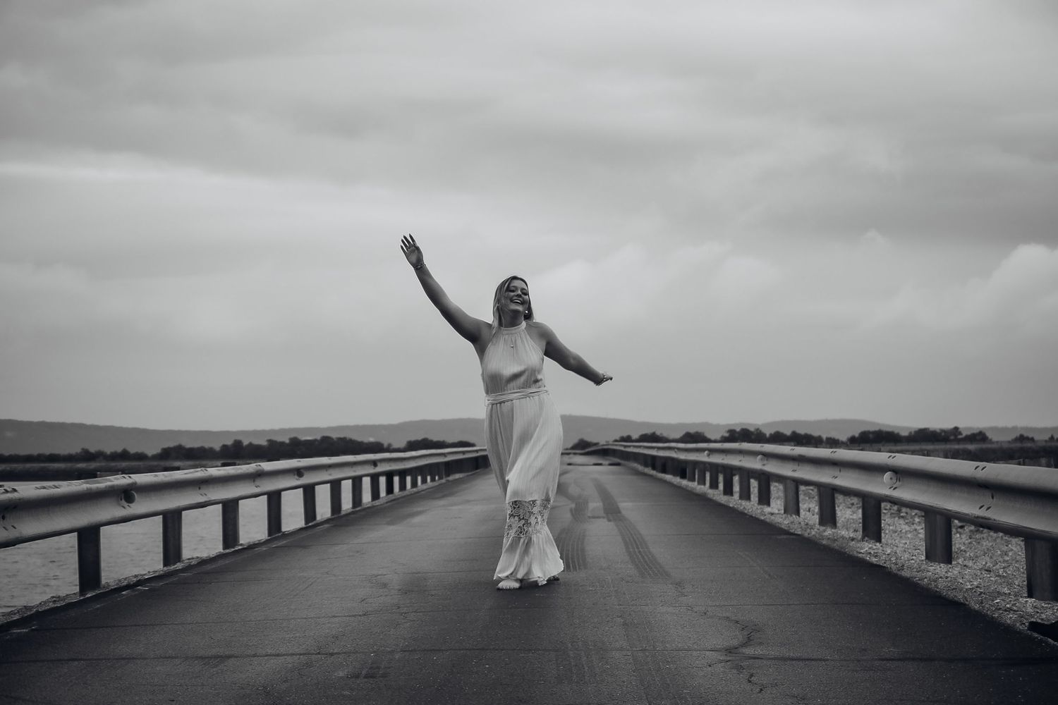 Woman in white dress dancing on an empty bridge under a cloudy sky in black and white.