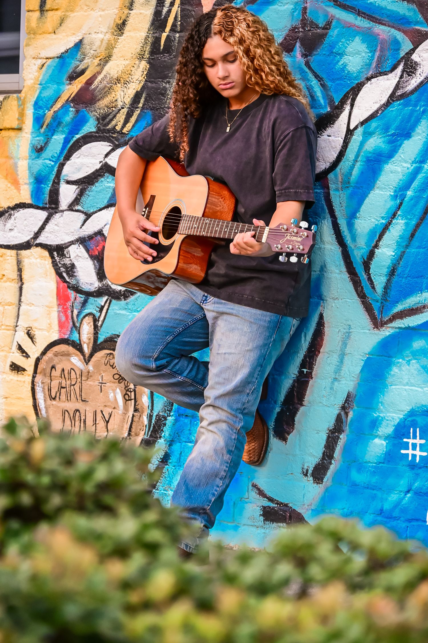 Young woman with curly hair playing acoustic guitar against a colorful graffiti wall outdoors.