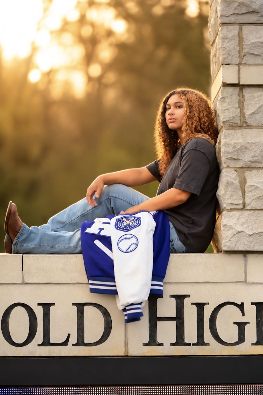 Teen girl in jeans and gray tee poses on Ringgold High School sign with blue varsity jacket at golden hour.