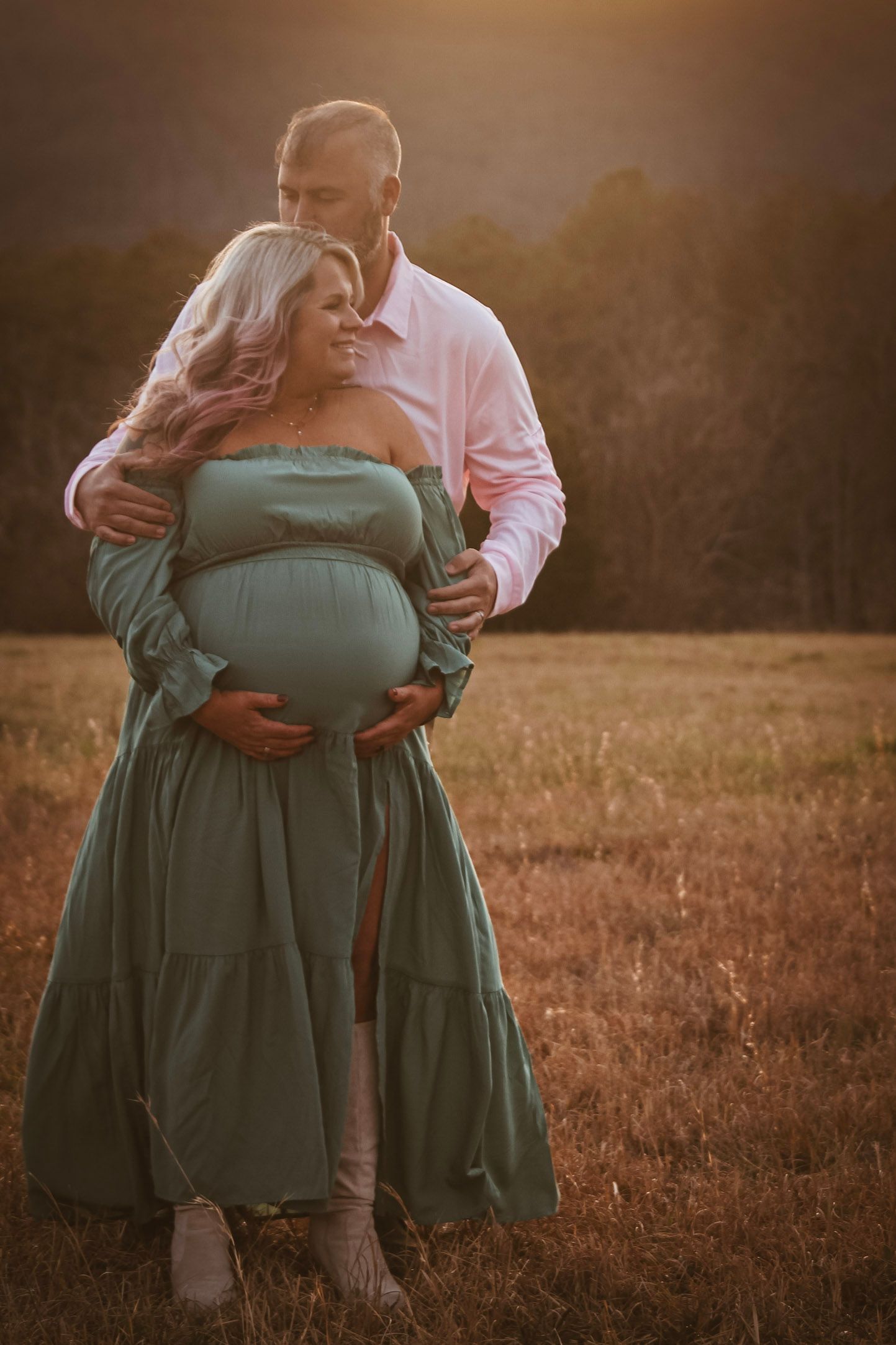 Pregnant couple embracing in a golden sunset field, woman in teal maternity gown.