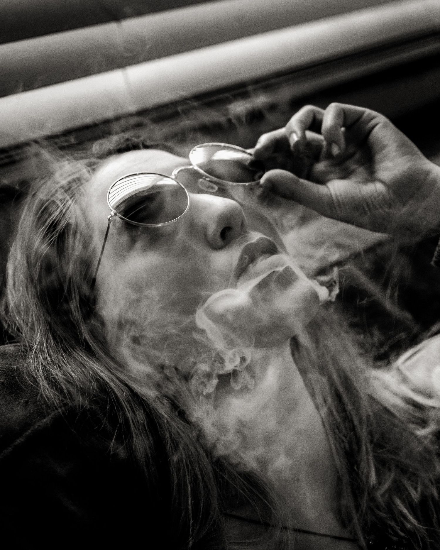Black and white photo of a woman with long hair and round sunglasses exhaling smoke, surrounded by dramatic swirling clouds.