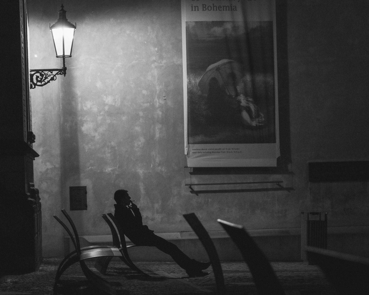 Black and white night photo of a solitary person sitting on a bench under a street lamp.