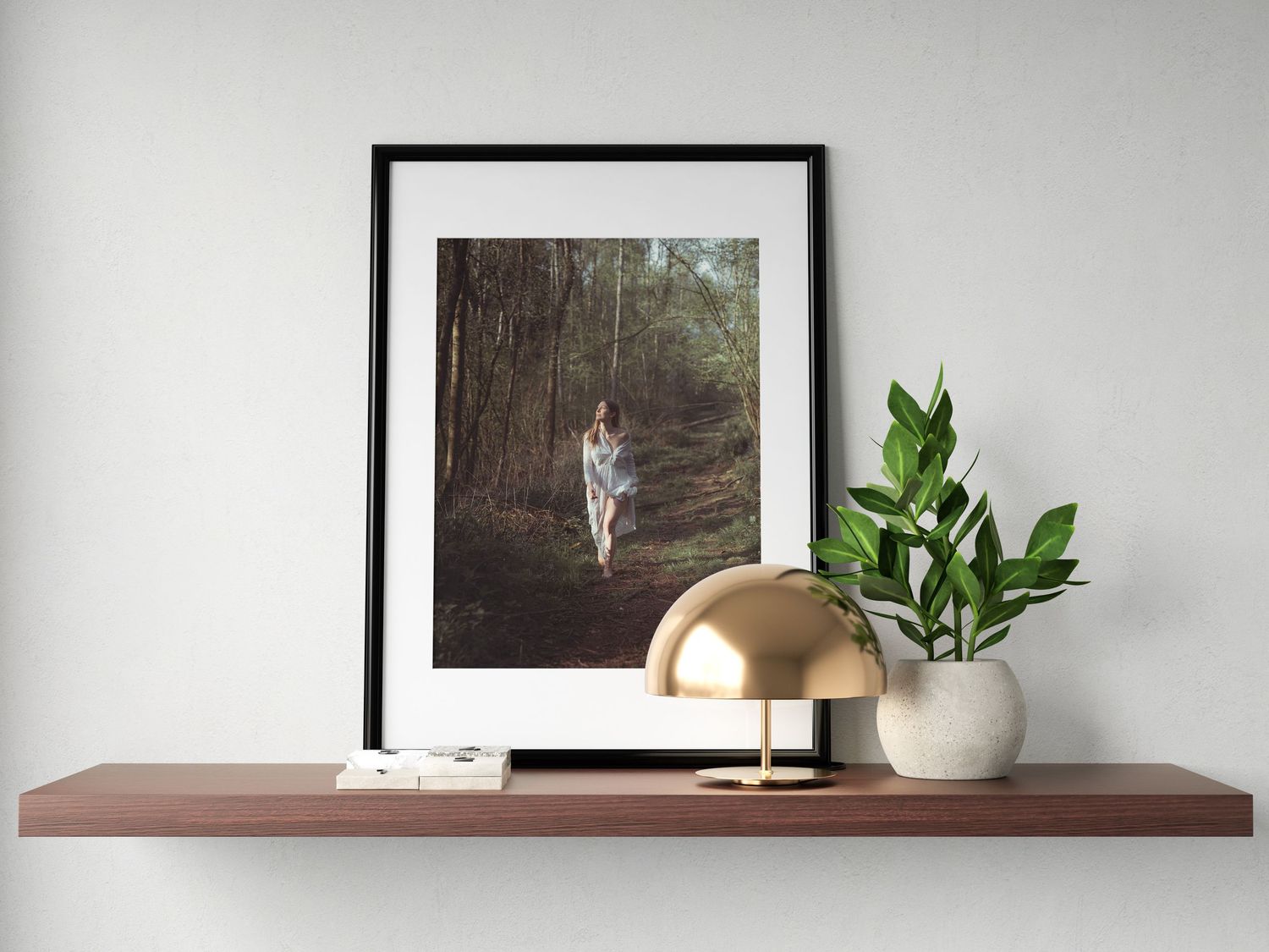 Framed forest swing photo on a walnut shelf with a gold dome lamp and white vase with green plant.