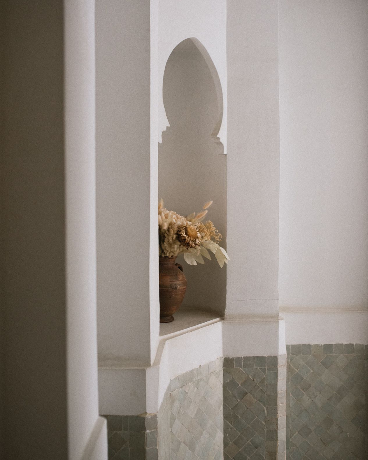 Dried flowers in a terracotta vase displayed in a Moorish arched white wall niche.