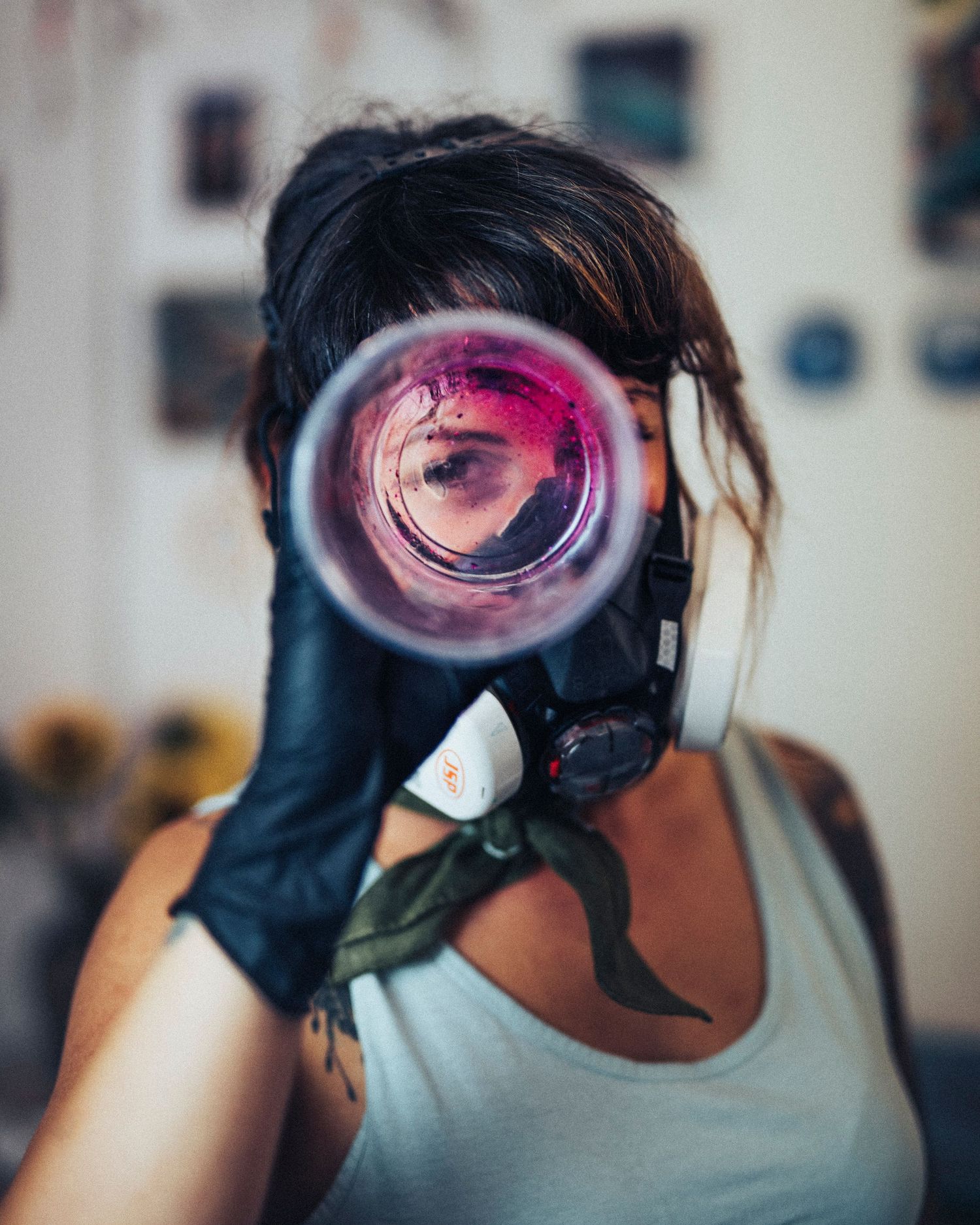 Woman in gas mask and black gloves holds pink-tinted lens up to her eye in a creative studio setting.