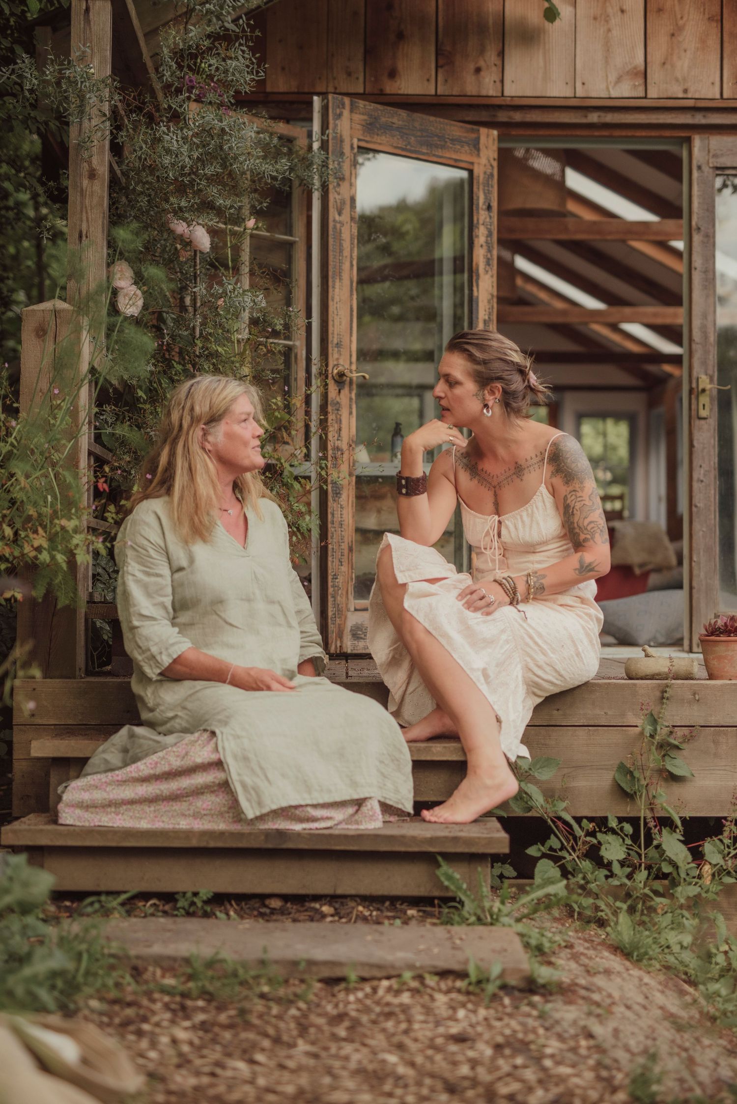 Two women in white dresses sit chatting on wooden steps of a rustic cabin surrounded by greenery.