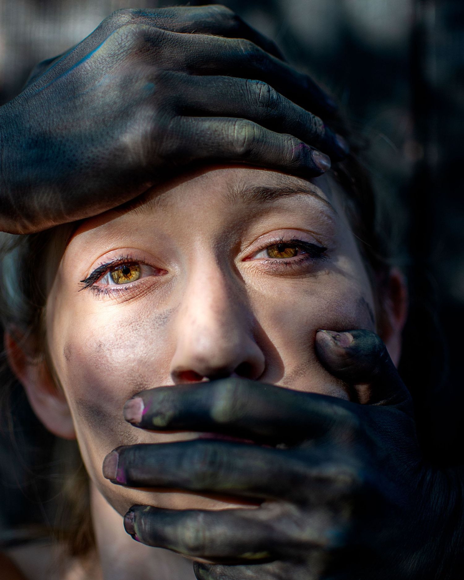 Close-up of a woman with golden eyes, dark hands covering her mouth and forehead in a dramatic artistic portrait.