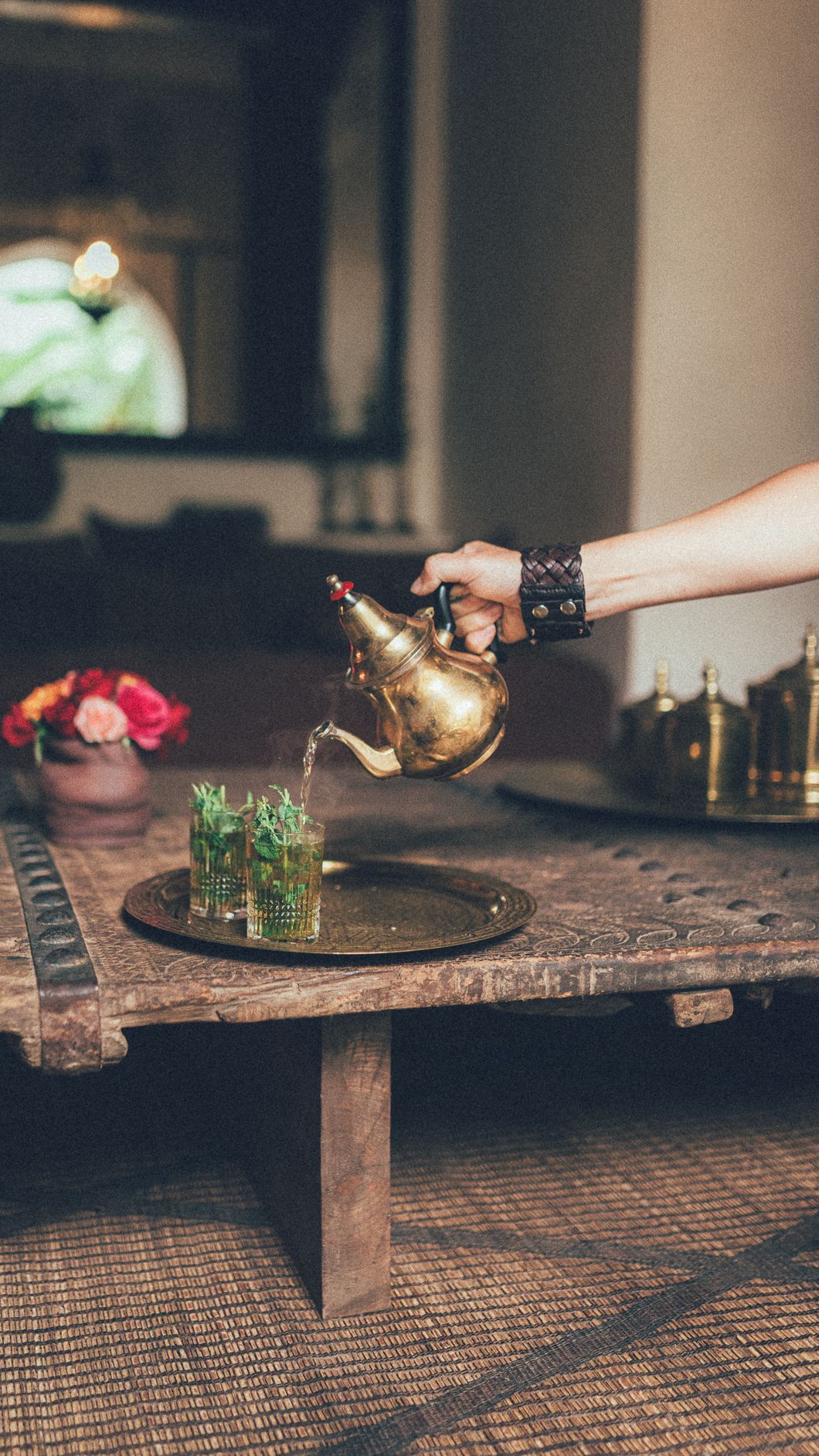 Hand pouring Moroccan mint tea from a gold teapot into glasses on a wooden tray.