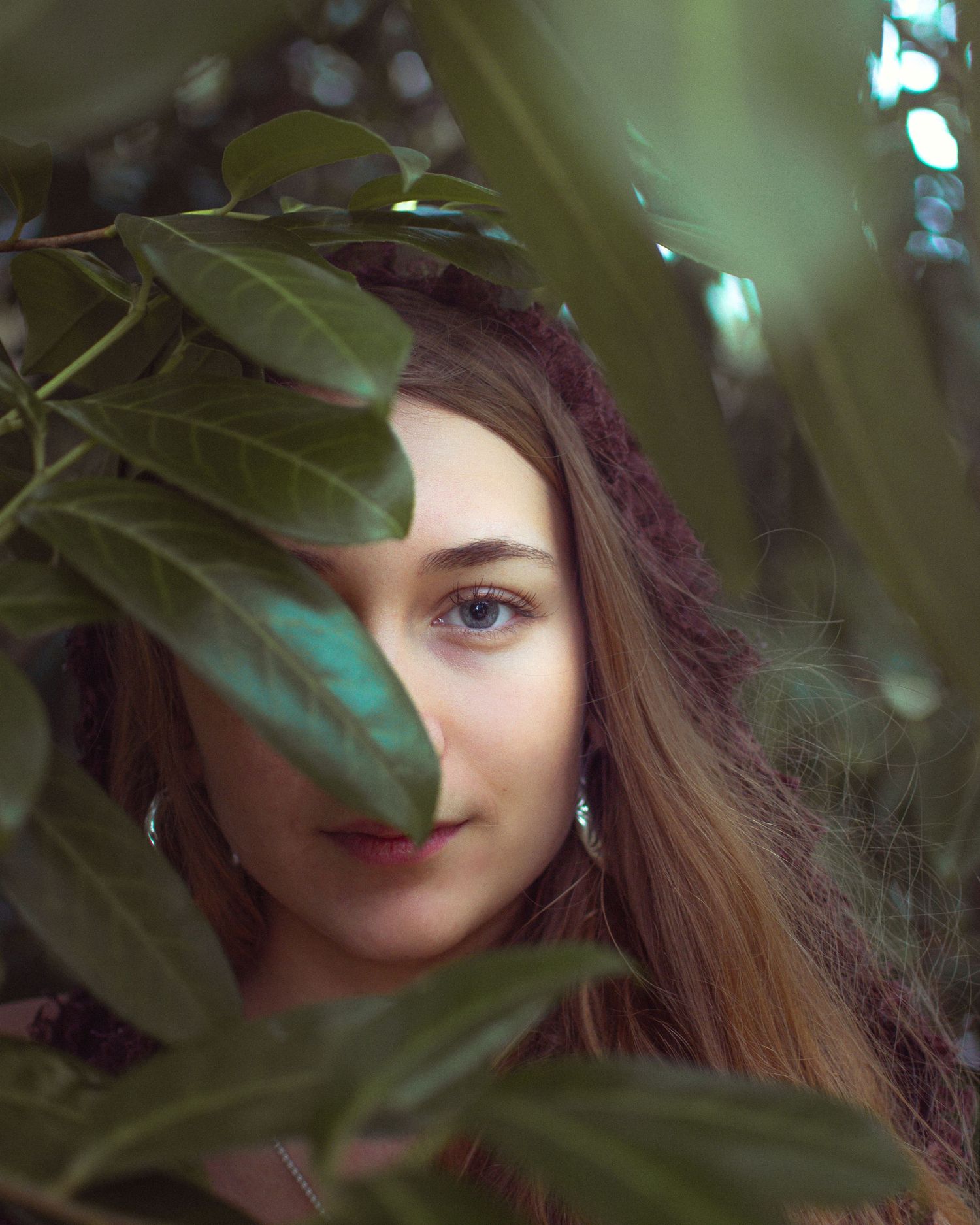 Young woman with long brown hair peering through lush green tropical leaves, one eye visible, wearing a knit hat.