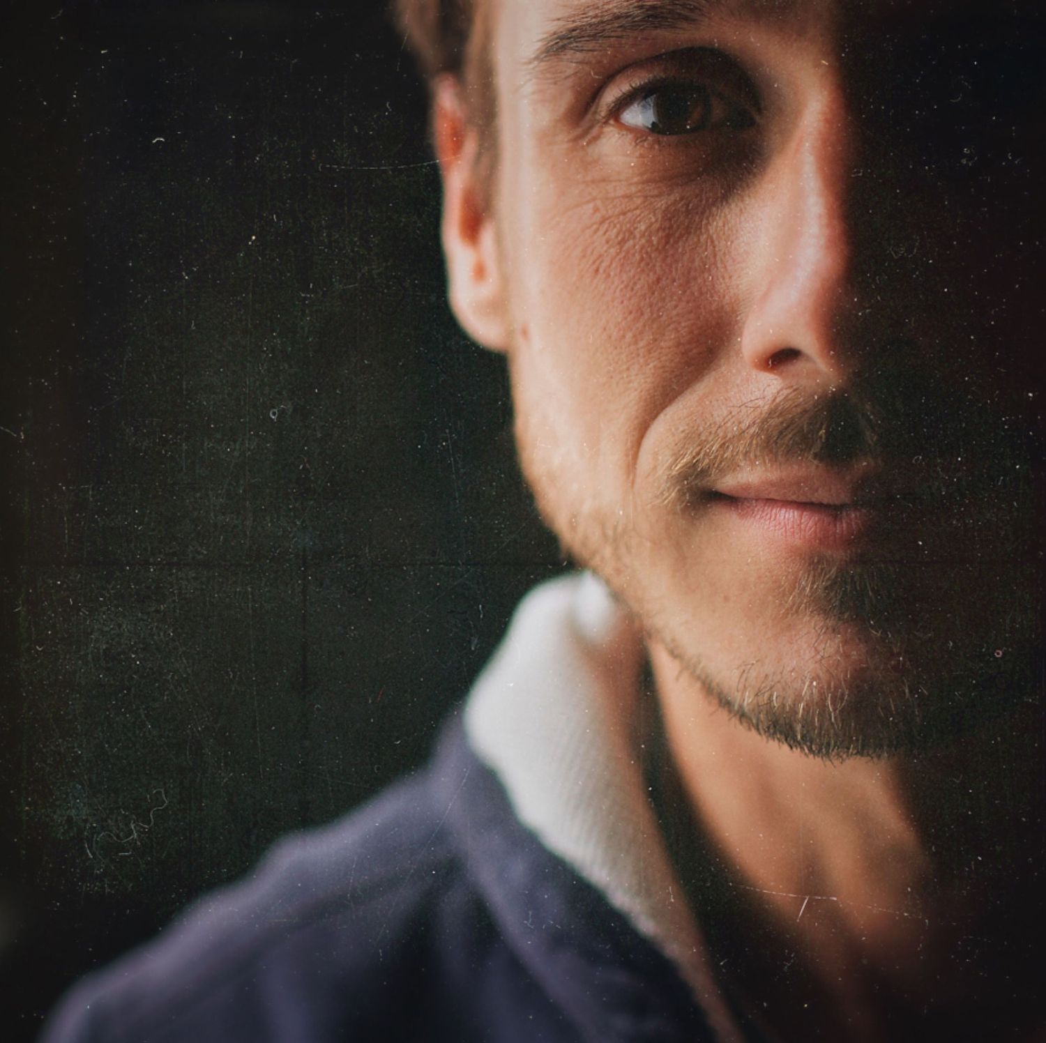 Close-up portrait of a man with stubble and a subtle smile against a dark textured background.