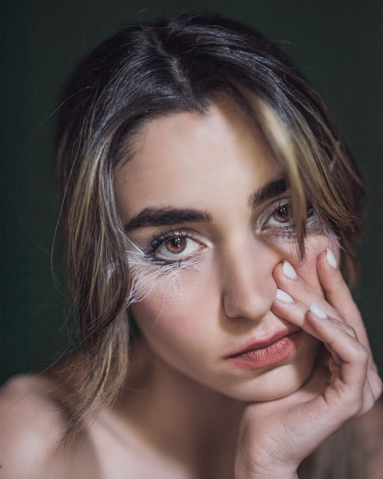 Close-up portrait of young woman with feather eye makeup resting hand against her cheek.