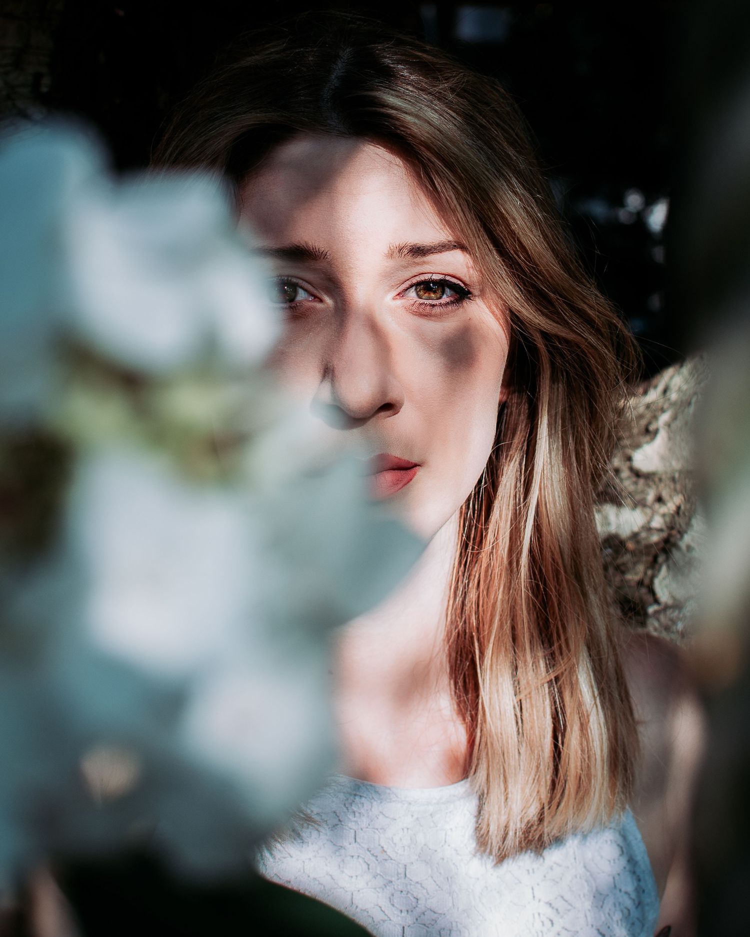 Young woman with brown hair gazes through white flowers in soft natural light, wearing a white lace top.
