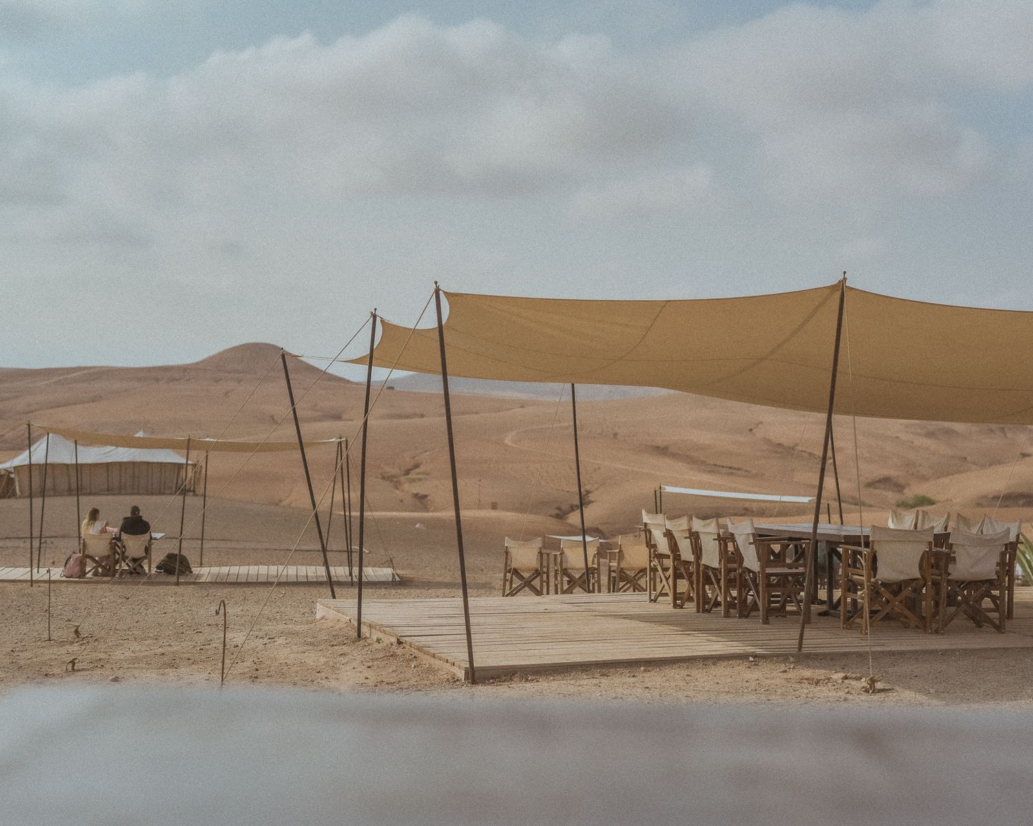 Desert dining setup with tan shade canopies and long table among sandy dunes.