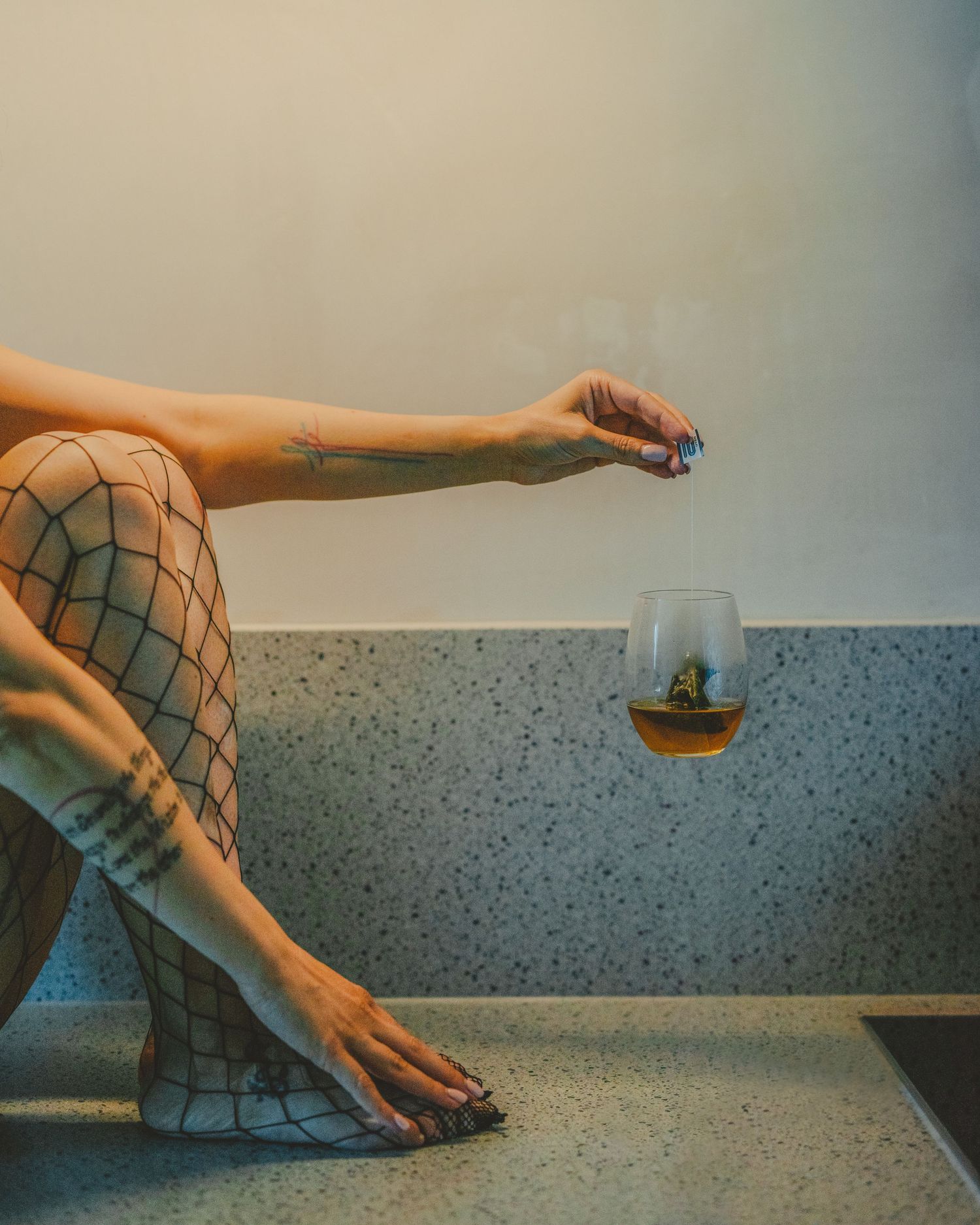 Woman in fishnet tights sitting on counter holding a stemless glass of whiskey with tattoos visible.