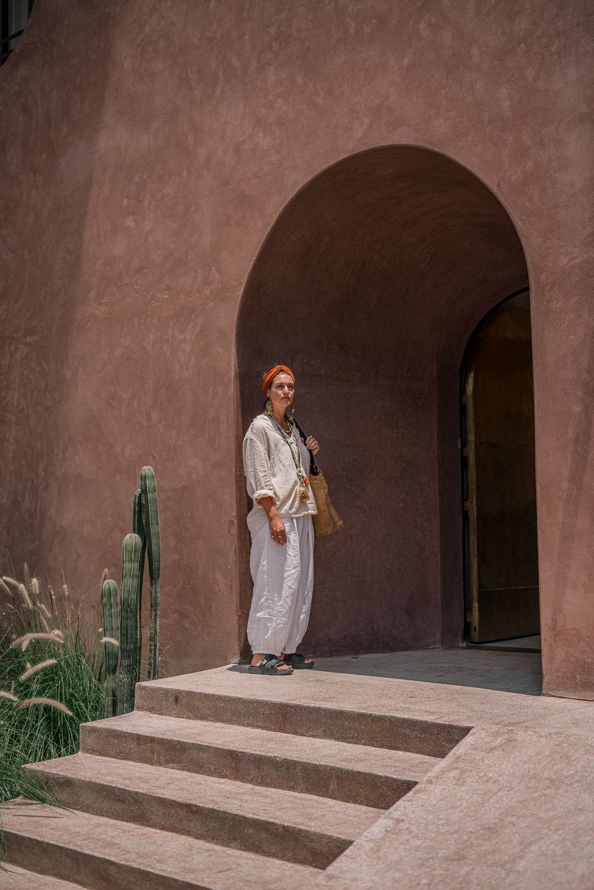 Woman in white linen outfit standing under adobe arch with cacti, desert resort architecture.