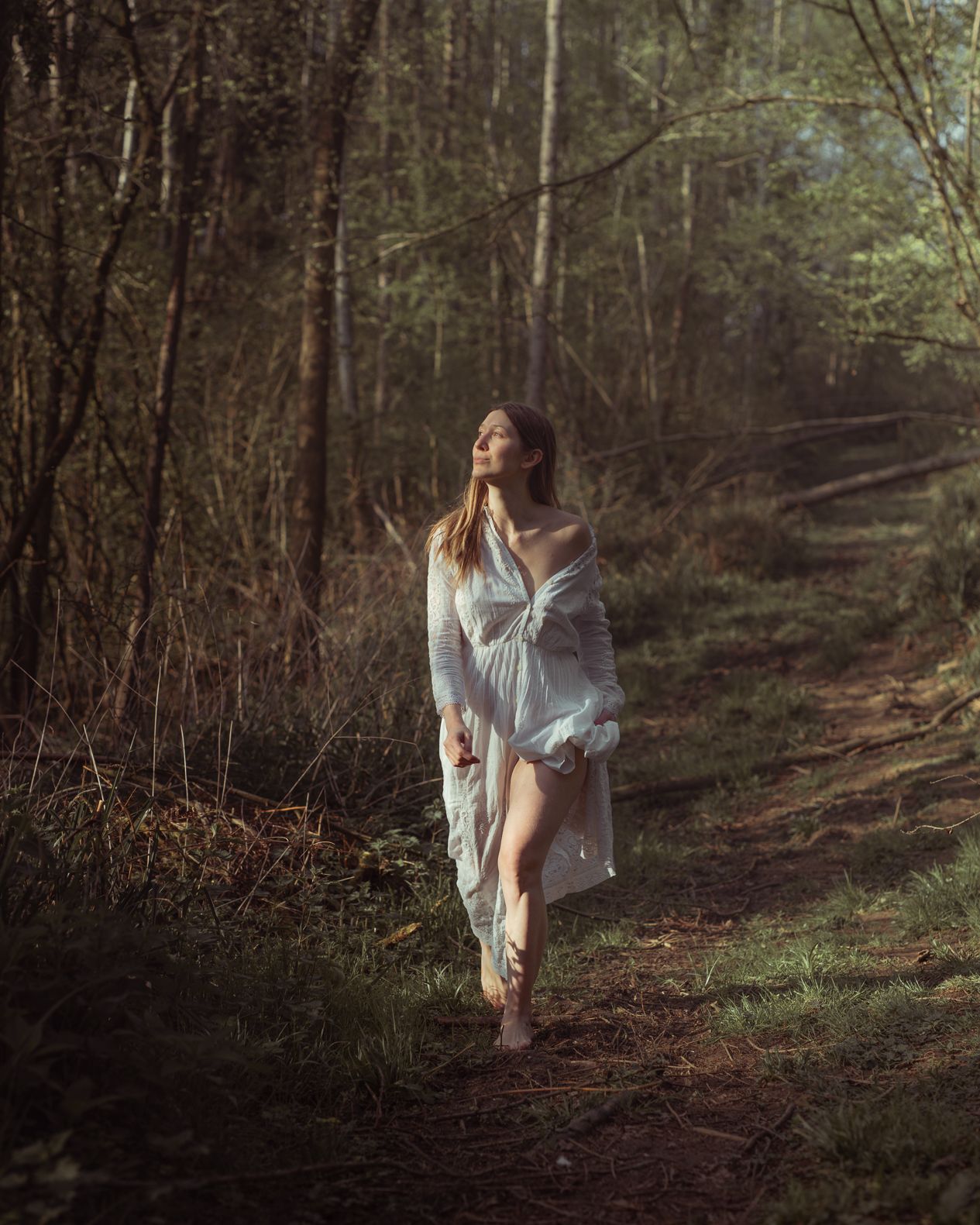 A woman in a flowing white dress walks barefoot along a sunlit forest path.