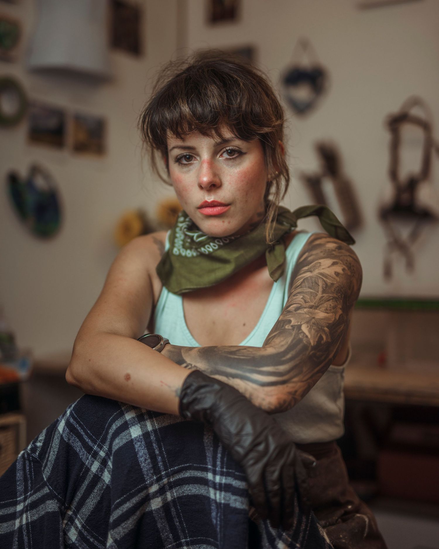 Tattooed woman with bangs wearing a green bandana and plaid blanket, sitting cross-armed in a rustic studio.