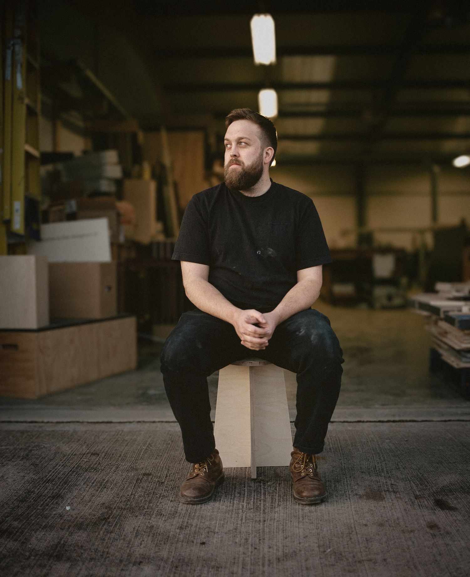Bearded man in black t-shirt sits on wooden box in industrial warehouse, wearing brown boots, hands clasped.