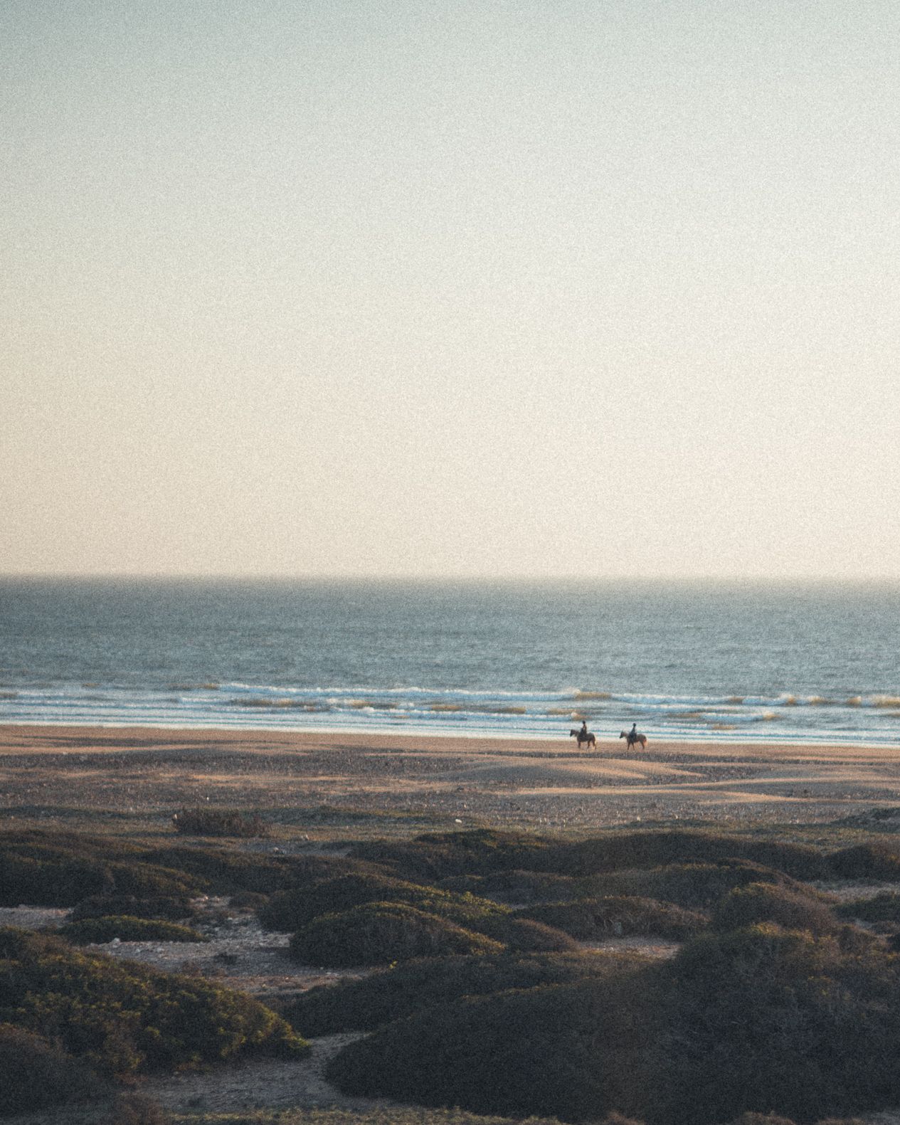 Two horse riders on a vast sandy beach at golden hour with calm ocean waves and dunes in the foreground.