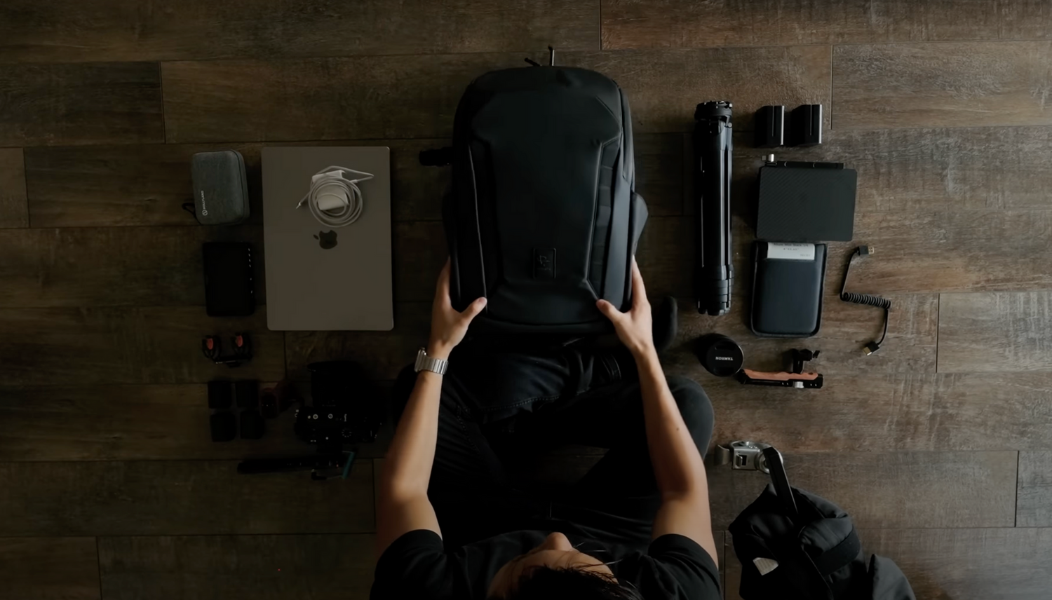 Top-down perspective of hands holding open a dark backpack with tech gear visible.