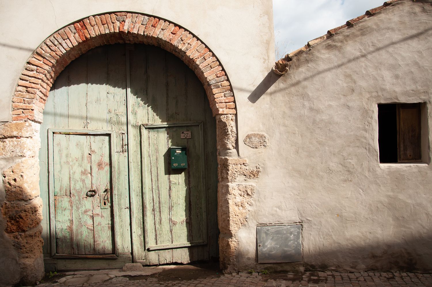 Rustic green wooden door set in a brick arch against a weathered stone wall in a cobblestone alley.