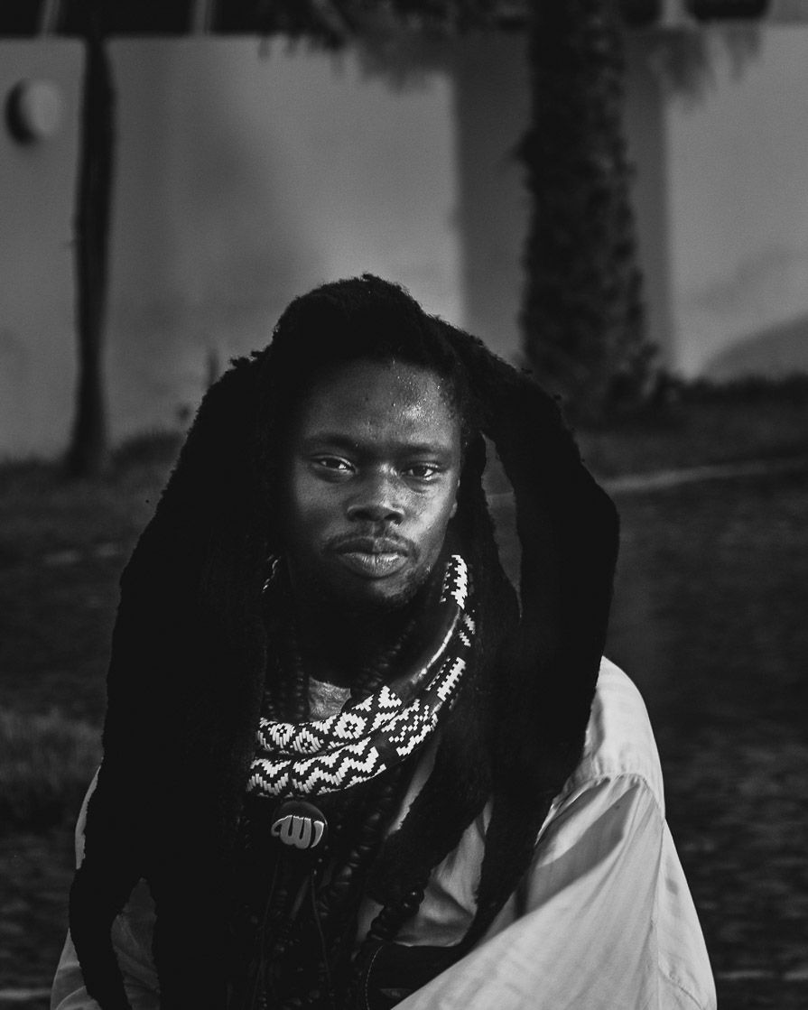 Black and white portrait of a man with dreadlocks and beaded necklace sitting outdoors at night.
