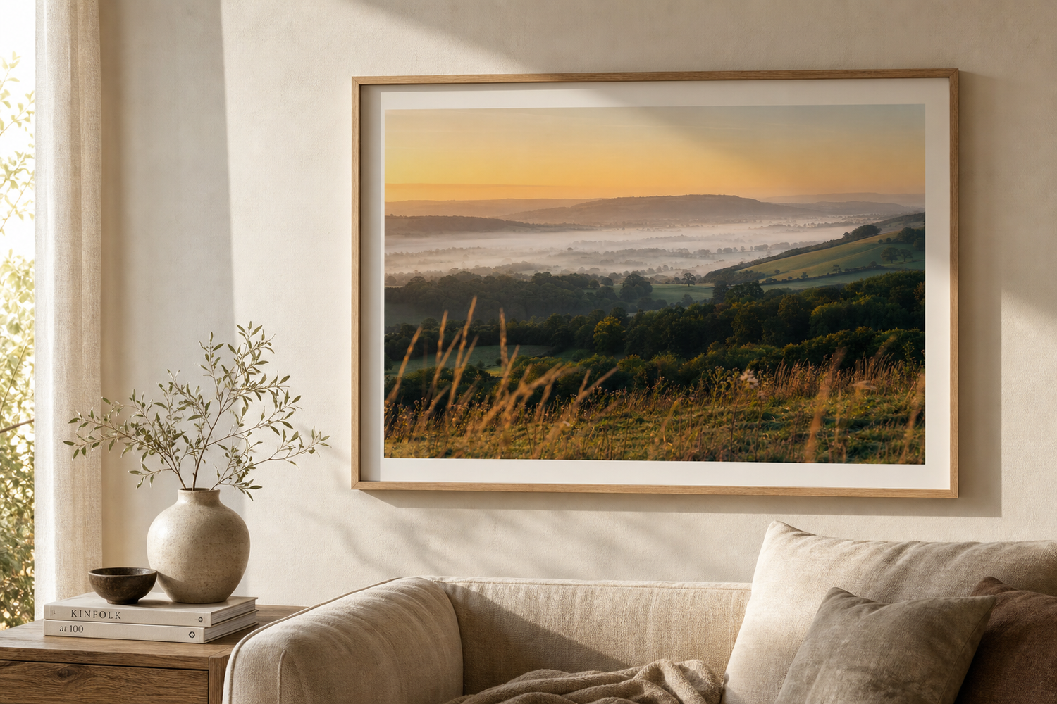 Framed landscape print of misty rolling hills at golden sunset hour hanging above a cozy neutral living room sofa.
