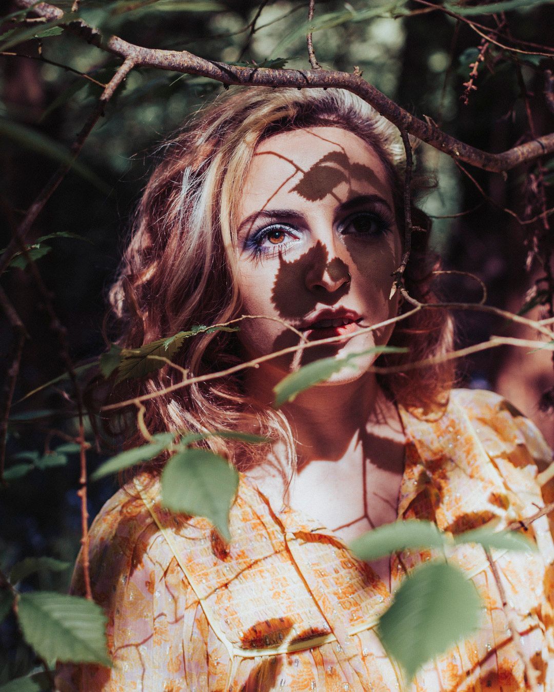 A young woman with blue eyes and blonde hair peers through forest branches, dappled shadows across her face, wearing a floral top.