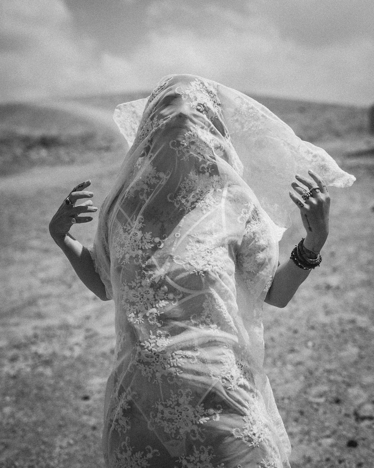 Black and white photo of a woman draped in flowing lace veil outdoors in a desert landscape.