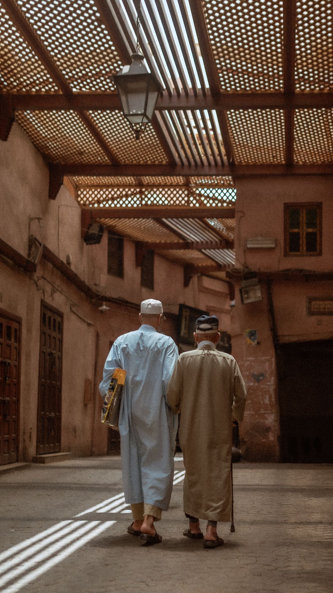 Two men in traditional robes walking through a sunlit Moroccan medina alleyway with ornate latticed ceiling.
