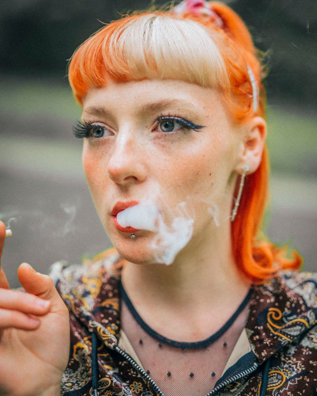 Young woman with vibrant orange and blonde hair exhaling smoke, wearing a paisley jacket and layered necklaces outdoors.