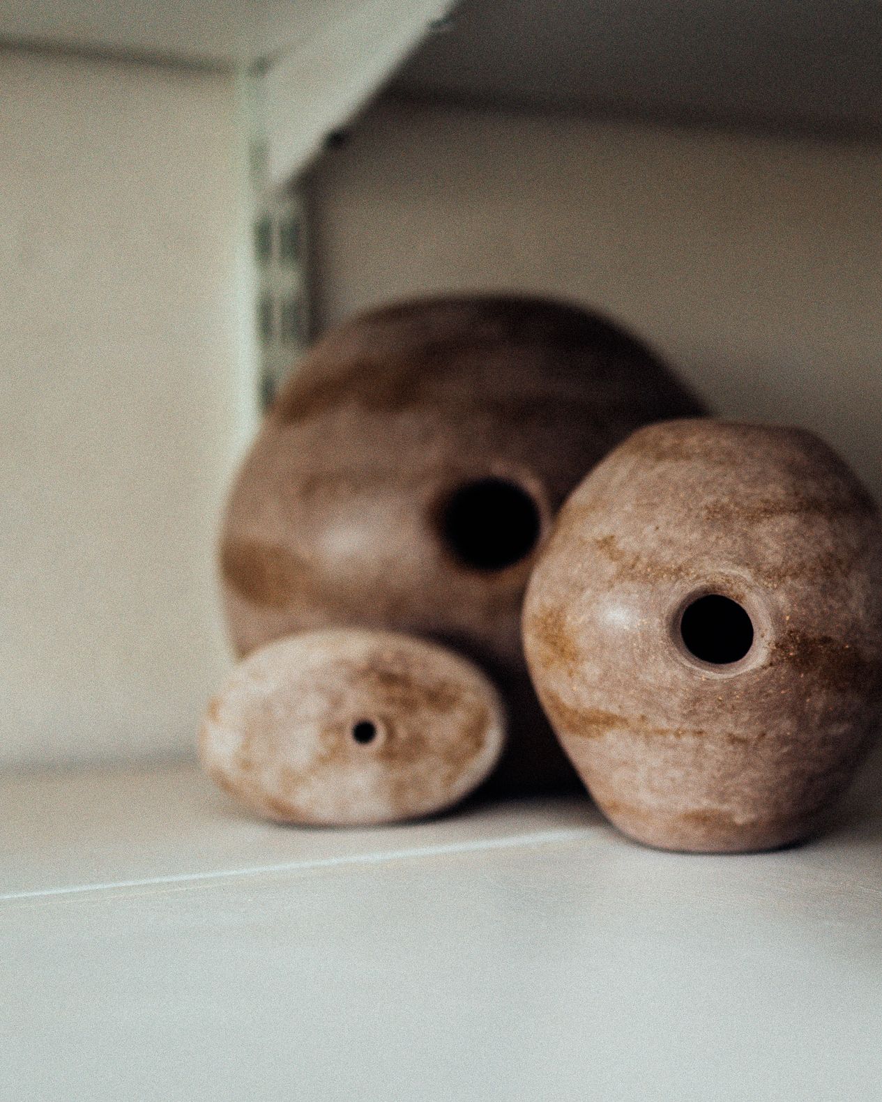 Three rustic brown ceramic vessels with small holes displayed on a white shelf.