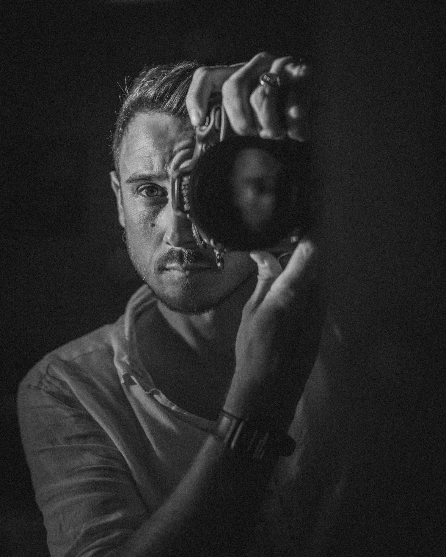 Black and white portrait of a man holding a camera up to his eye, wearing a watch and white shirt.